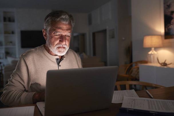Senior uomo ispanico controllare le sue finanze online a casa utilizzando un computer portatile di notte
 - Foto, immagini