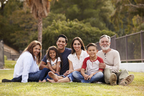 Three generation Hispanic family sitting on the grass in the park smiling to camera - Photo, Image