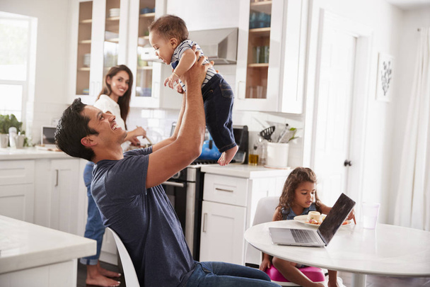 Young Hispanic family in their kitchen, dad lifting baby in the air, mum cooking at hob - Foto, afbeelding
