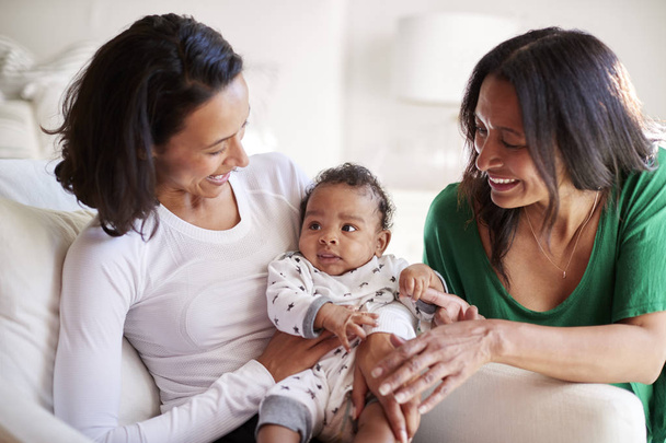 Millennial mixed race mother sitting in an armchair holding her three month old baby son, her mother kneeling beside them - Photo, image