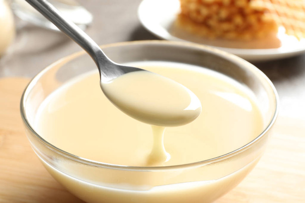Spoon of pouring condensed milk over bowl on table, closeup. Dairy products - Fotografie, Obrázek