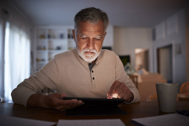 Senior Hispanic man sitting at a table reading an e book at home in the evening, close up - Foto, imagen