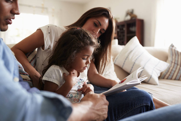 Hispanic couple and their young daughter sitting on the sofa reading a book together at home - Photo, Image