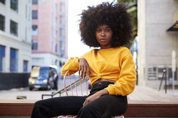Young black woman with afro hair and yellow top sitting on a chair in the street looking to camera - Fotoğraf, Görsel