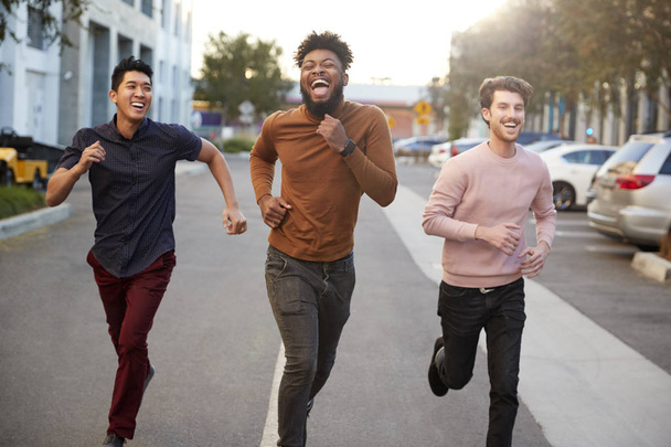 Three hip young adult male friends running for fun in a city road, front view, three quarter length - Foto, immagini