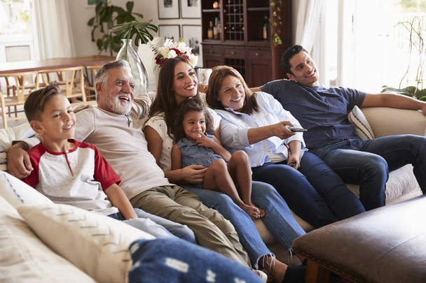 Три покоління латиноамериканського сімейства сидять на дивані дивляться телевізор, бабуся з допомогою пульта дистанційного керування - Фото, зображення