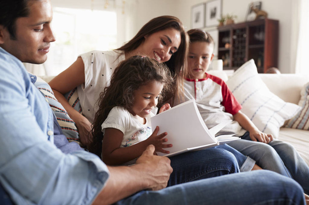 Joven familia hispana de cuatro personas sentadas en el sofá leyendo el libro juntas en su sala de estar
 - Foto, Imagen