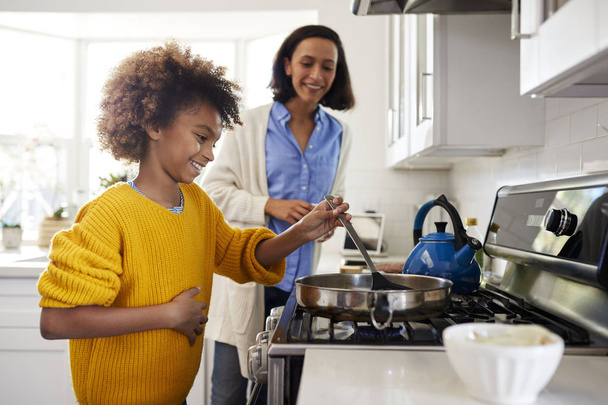 Pre-teen girl standing at hob in the kitchen using spatula and frying pan, preparing food with her mother, side view - Photo, image