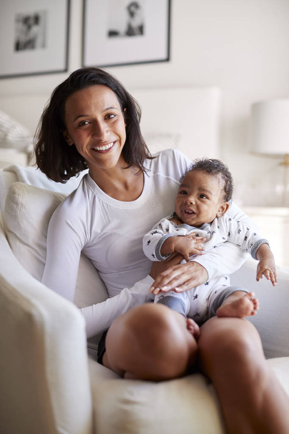 glücklich gemischte Rasse junge erwachsene Mutter sitzt auf einem Sessel hält ihren drei Monate alten kleinen Sohn, lächelt in die Kamera, vertikal - Foto, Bild