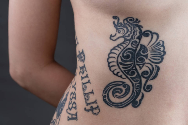 A tattoo of a black tribal style seahorse with children's names wrapping around a woman's ribs. - Foto, Imagem