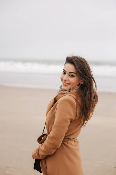 Portrait of beautiful woman walkink along the coast near the north sea. Elegant lady in brown coat with fashion make up stand in front of the sea - Фото, изображение