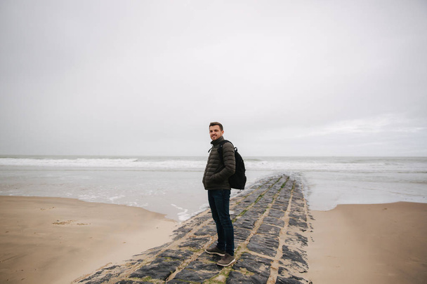 Male tourist walkin along the North Sea. Man with backpack stand on the coast in rainy weather. Beautiful sea - Valokuva, kuva