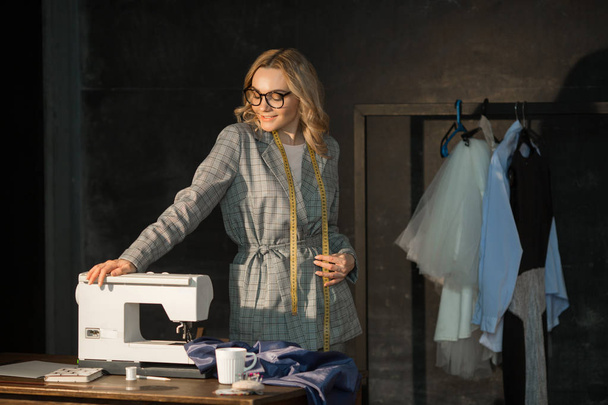 beautiful young girl seamstress in glasses in a jacket at work stands near the sewing machine in a dark room - 写真・画像