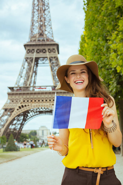 smiling trendy tourist woman in yellow blouse and hat showing French flag against clear view of the Eiffel Tower in Paris, France. - Фото, изображение