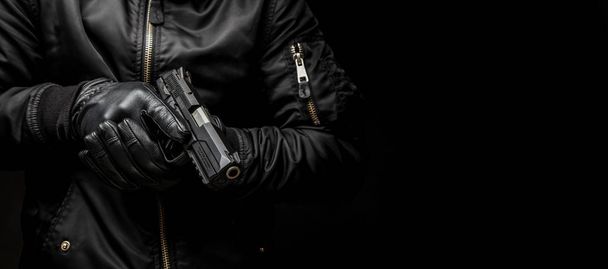 a man in a black jacket and black gloves holding a gun on a dark back - Foto, Bild