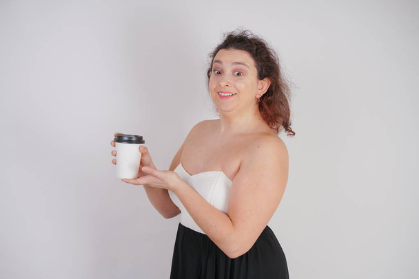 feminine woman with plus size body in a fashionable dress holding a paper Cup of coffee and posing on a white background in the Studio - Фото, изображение