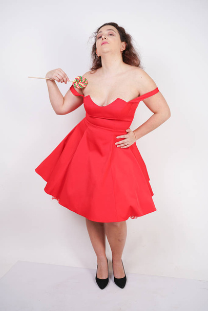 całkiem pulchna dziewczyna moda ubrana czerwona sukienka i pozowanie z smaczne kolorowe lizak na białym tle Studio - Zdjęcie, obraz