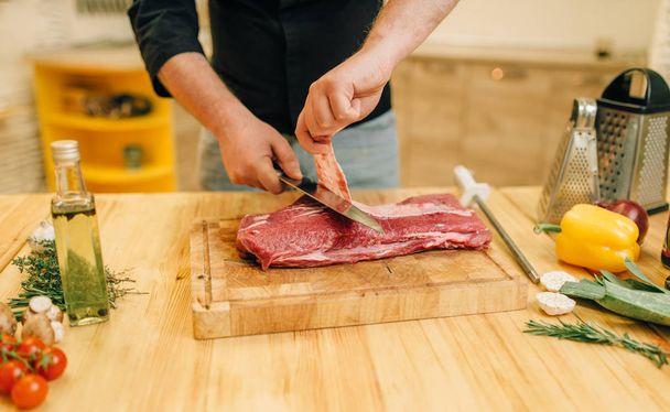 Male person with knife cuts raw meat on wooden board, kitchen interior on background. Chef cooking tenderloin with vegetables, spices and herbs - Photo, Image