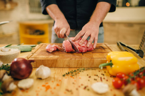 Male person with knife cuts raw meat into slices, closeup, kitchen interior on background. Chef cooking tenderloin with vegetables, spices and herbs - Fotó, kép