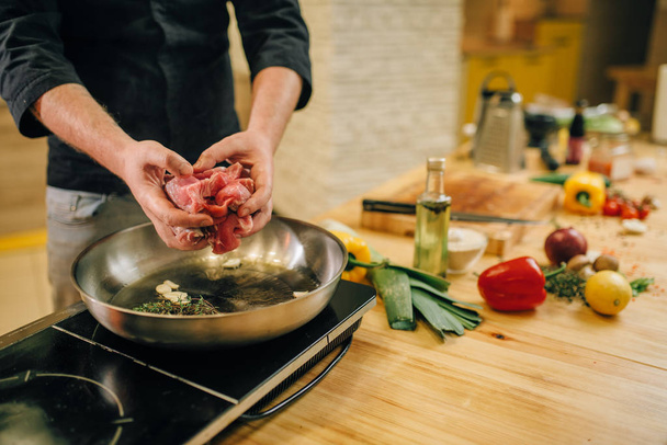 男性は台所の鍋でハーブや調味料で肉を調理します。テーブルの電気ストーブの上でゆでた豚肉を準備する男 - 写真・画像