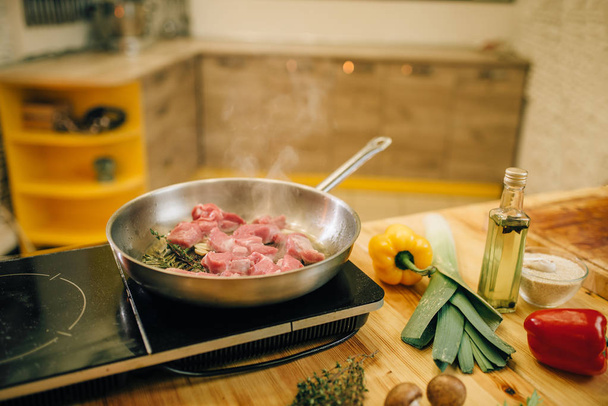Filet de viande rôti dans une poêle à frire sur cuisinière électrique, légumes sur planche de bois sur fond, personne. Boeuf, assaisonnements et épices avec des herbes sur le comptoir
 - Photo, image
