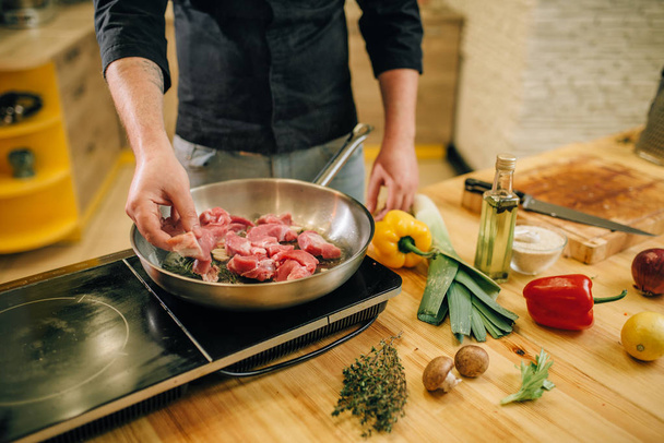 Férfi személy főzés húst gyógynövények és fűszerek egy serpenyőben a konyhába. Ember készül főtt sertéshús asztali villanytűzhely - Fotó, kép
