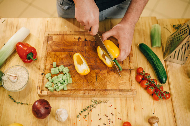 Male chef with knife cuts yellow pepper on wooden board, top view. Man cutting vegetables, fresh salad cooking, kitchen interior on background - Photo, Image