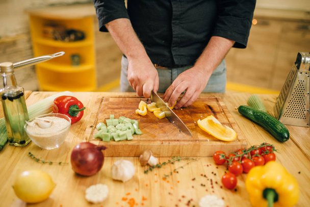 Chef hands with knife cuts yellow pepper on wooden board closeup. Man cutting vegetables, fresh salad cooking, kitchen interior on background. Male person chopping ingredients for lettuce - Фото, зображення