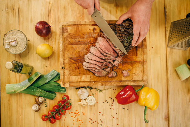 Male chef hands with knife cuts roasted meat on slices, top view. Man preparing beef with vegetables on countertop - Photo, image