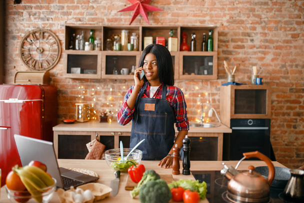 Musta nainen esiliinassa puhuu matkapuhelimella keittiössä. Afrikkalainen nainen valmistelee kasvissalaattia kotona, etninen tyttö ruoanlaitto terveellinen aamiainen
 - Valokuva, kuva
