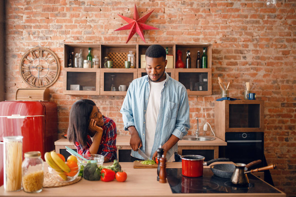 Schwarzer Mann kocht Frühstück in der Küche, Frau trinkt Kaffee. Afrikanisches Paar bereitet Gemüsesalat zu Hause zu - Foto, Bild