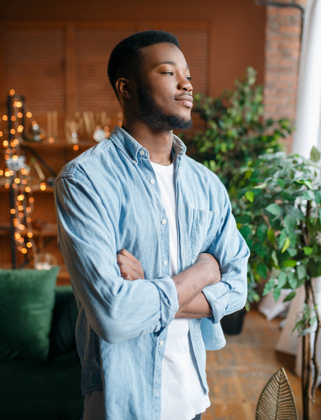 Zwarte man met een sluwe blik in de woonkamer, ontspanning thuis. Jonge Afro-Amerikaanse mannelijke persoon die in zijn huis staat - Foto, afbeelding
