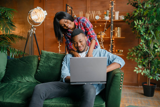 Μαύρο ζευγάρι με φορητό υπολογιστή που διασκεδάζει στον καναπέ στο σπίτι. Ευτυχισμένη αφρικανική αγάπη ζευγάρι λειάζει στο σπίτι τους, χαρούμενη οικογένεια χαλάρωση στο σπίτι - Φωτογραφία, εικόνα