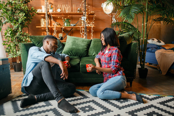 Черная пара сидит на полу и пьет кофе против дивана дома. Счастливые африканцы любят пару досуга в их доме, веселые семьи расслабляются вместе утром
 - Фото, изображение