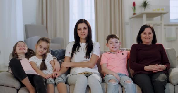 白い笑顔を持つカリスマ的な女性と彼女の3人の子供たちは、リビングルームのソファに座っている間、おばあちゃんと一緒にテレビを見て時間を過ごす. - 映像、動画