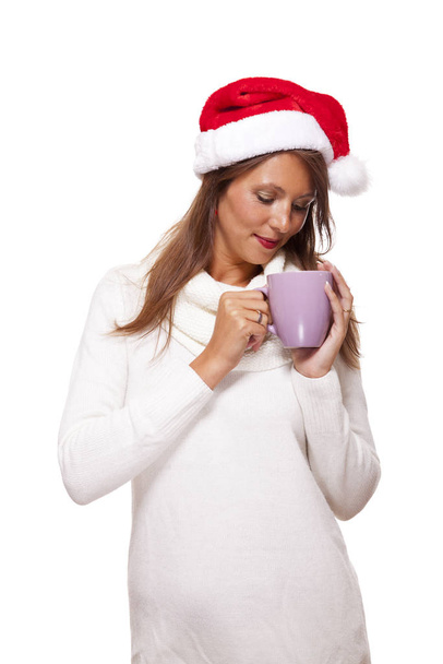 Cold attractive young woman with a cute smile in a festive red Santa hat sipping a hot mug of coffee that she is cradling in her hands to warm up in the winter weather, on white - Fotoğraf, Görsel