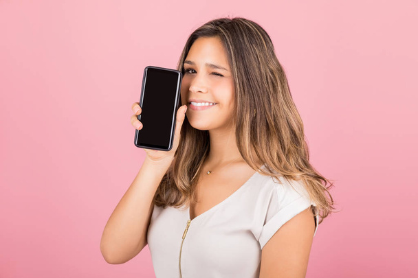 Portrait of woman winking while holding cell phone with blank screen in studio - Foto, Imagen