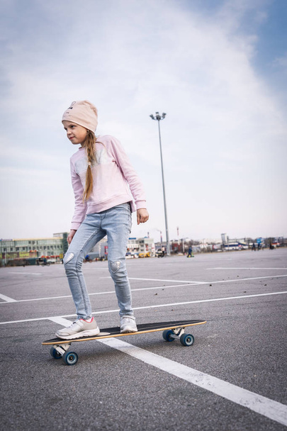 A teenager girl in a pink hat and jeans is rolling on a longboard in the city. - Photo, image