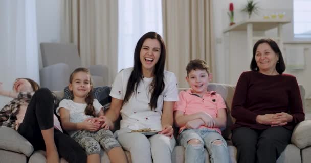 Happy smiling charismatic kids with their mother and granny watching a comedy movie on the TV in front of the camera while sitting on the sofa - Metraje, vídeo