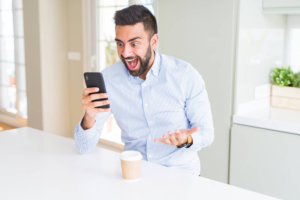 Hombre de negocios hispano guapo bebiendo café y usando un teléfono inteligente muy feliz y emocionado, expresión ganadora celebrando la victoria gritando con una gran sonrisa y levantando las manos
 - Foto, Imagen