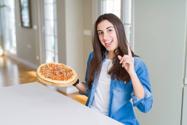 Belle jeune femme mangeant une pizza savoureuse maison à la cuisine surprise par une idée ou une question pointant du doigt avec un visage heureux, numéro un
 - Photo, image