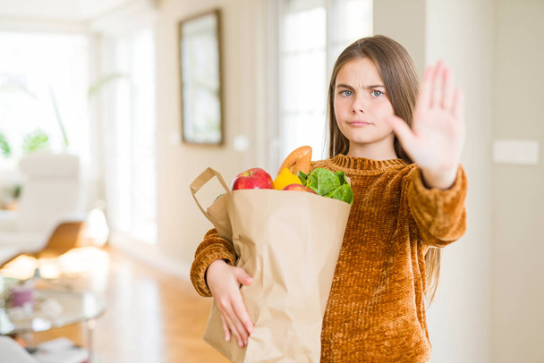 Krásná mladá dívka drží papírový sáček čerstvých potravin s otevřenou rukou dělá stopku s vážnou a sebevědomý výraz, gesto obrany - Fotografie, Obrázek