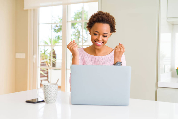 Młody African American kobieta pracy za pomocą komputera laptop obchodzi zaskoczony i zdumiony sukcesem z ramionami podniesione i otwarte oczy. Koncepcja zwycięzcy. - Zdjęcie, obraz