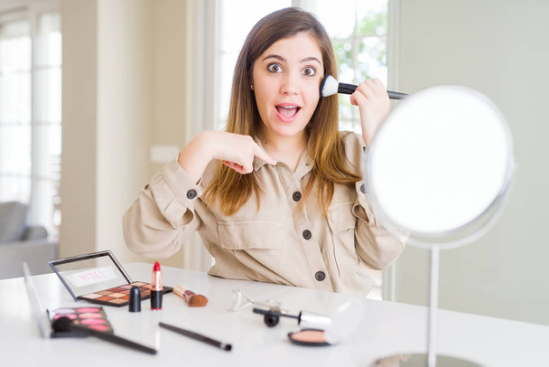 Piękna młoda kobieta za pomocą makijażu kosmetyki stosując kolor za pomocą pędzla z niespodzianką twarz palcem wskazującym do siebie - Zdjęcie, obraz