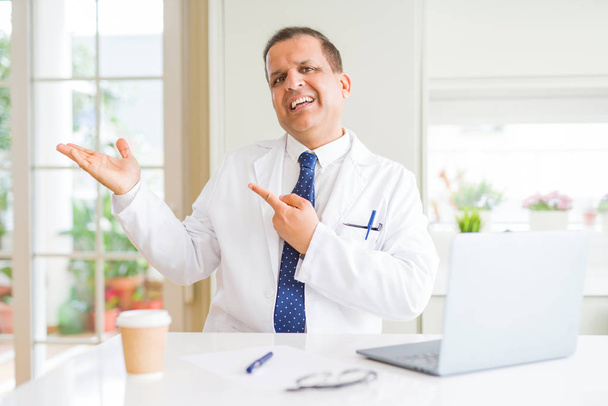 Muž středního věku doktor s bílým zdravotnickým kabátem, který pracuje s notebookem na klinice a s kamerou se usmívá, když se prezentuje s rukou a ukazuje prstem. - Fotografie, Obrázek