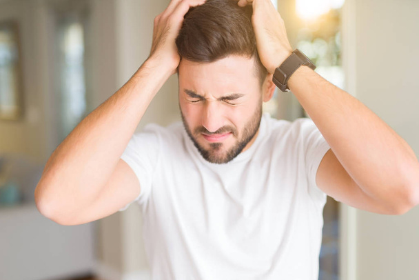 Young handsome man wearing casual white t-shirt at home suffering from headache desperate and stressed because pain and migraine. Hands on head. - Photo, Image