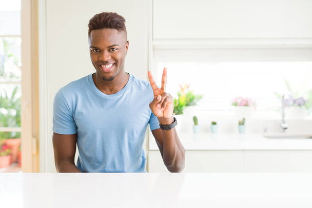 自宅でカジュアルなTシャツを着たハンサムなアフリカ系アメリカ人男性が自信を持って幸せに微笑みながら2番を指差す. - 写真・画像
