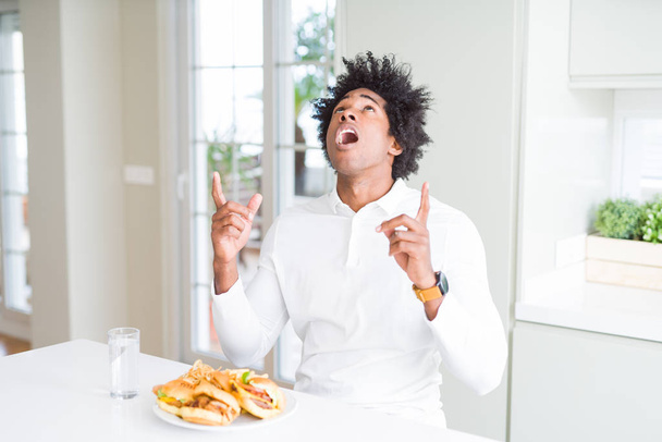Hombre afroamericano hambriento comiendo hamburguesa para el almuerzo asombrado y sorprendido mirando hacia arriba y señalando con los dedos y los brazos levantados
. - Foto, Imagen