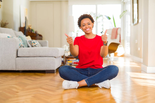junge schöne afrikanisch-amerikanische Frau sitzt auf dem Boden zu Hause Erfolgszeichen tun positive Geste mit der Hand, Daumen nach oben lächelnd und glücklich. Blick in die Kamera mit heiterem Gesichtsausdruck, Siegergeste. - Foto, Bild