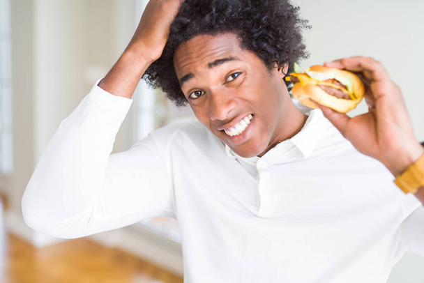 Uomo afroamericano affamato mangiare hamburger a pranzo stressato con mano sulla testa, scioccato con vergogna e faccia a sorpresa, arrabbiato e frustrato. Paura e sconvolto per errore
. - Foto, immagini